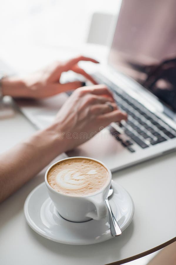Cup of hot coffee or cappuccino with a pattern. Blurred hands are typing on keyboard of a laptop in the background. Cup of hot coffee or cappuccino with a royalty free stock photography