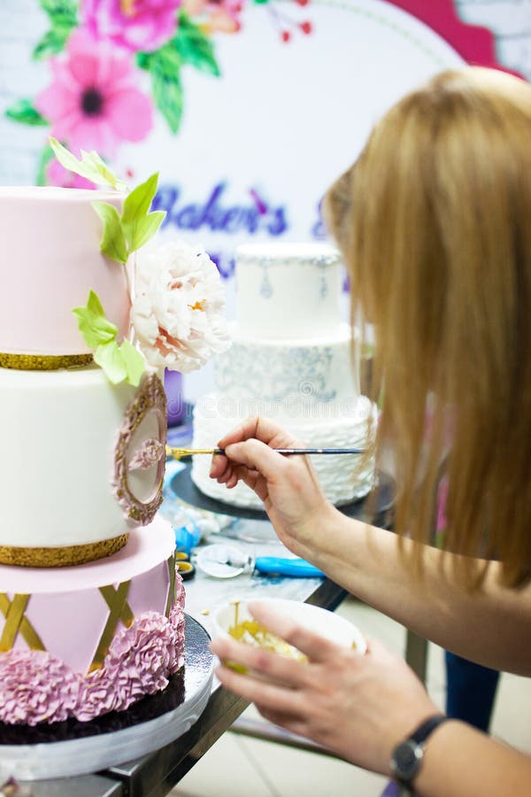 Decorating tiered mastic weeding cake with flowers and colouring details by brush. Young lady decorating tiered mastic weeding cake with flowers and colouring stock photos