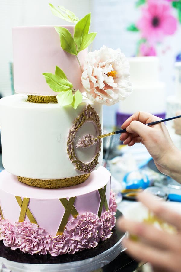 Decorating tiered mastic weeding cake with flowers and colouring details by brush. Young lady decorating tiered mastic weeding cake with flowers and colouring stock image
