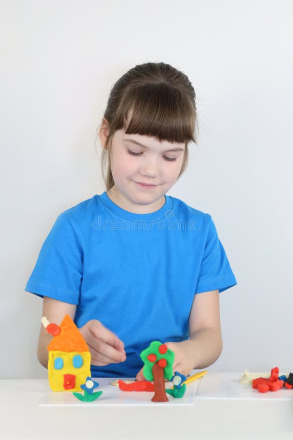 Smiling pretty girl molds toys from plasticine on table. In white room stock photo