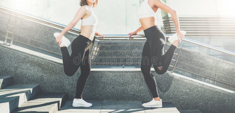 Two young women, girls runners are doing stretching exercises before running. Workout, training, couching on city street. Sunny summer day. Two young smiling stock photos